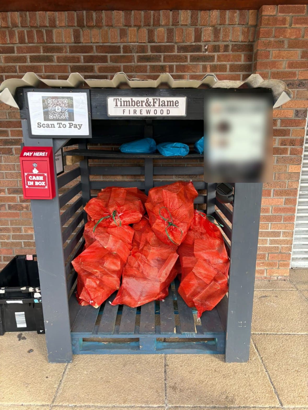 The Timber & Flame honesty stand: a slatted timber shelter stacked with red net bags of firewood, with a price board and cash box